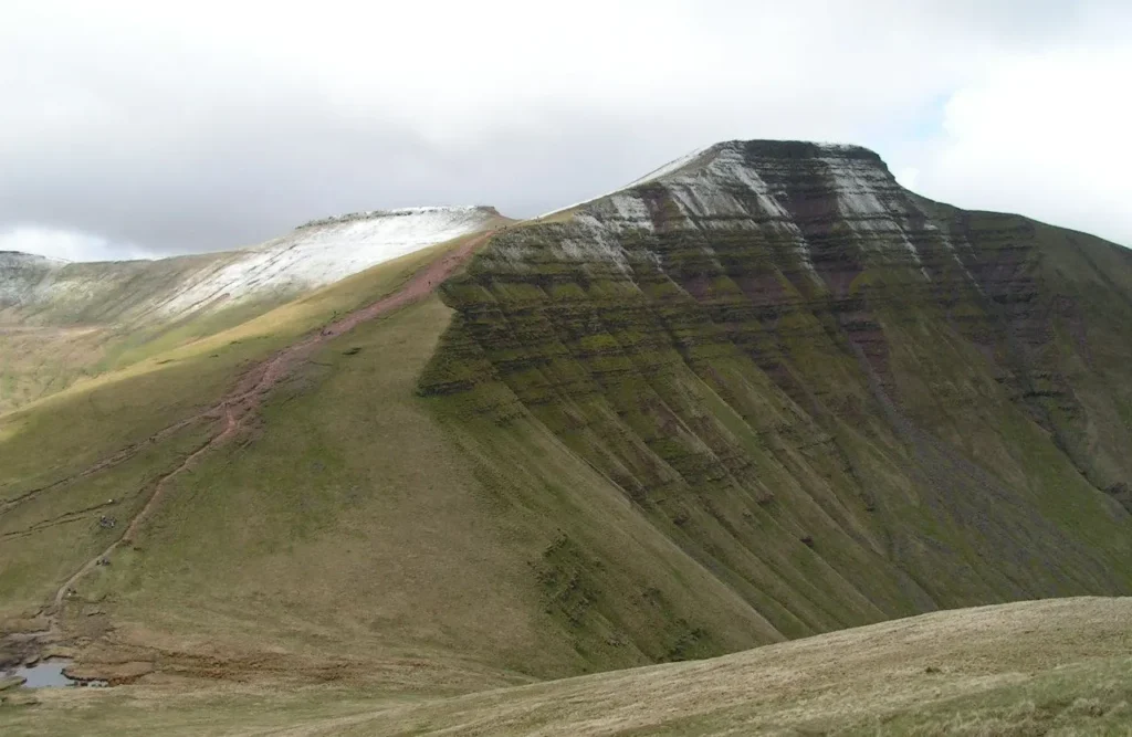 A striking view of Pen Y Fan, the highest peak in South Wales at 2,907 feet, partially dusted with snow near its summit. The mountain’s rugged, grassy slopes contrast against an overcast sky, showcasing the dramatic landscape of the Brecon Beacons National Park.