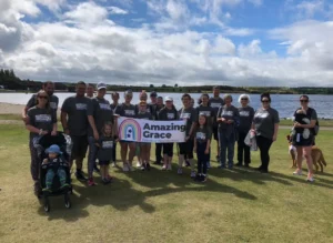 A group of people standing together, smiling and holding the Amazing Grace banner during a fundraising event. The backdrop features a serene lake, with clear blue skies and soft white clouds, creating a peaceful and uplifting atmosphere.