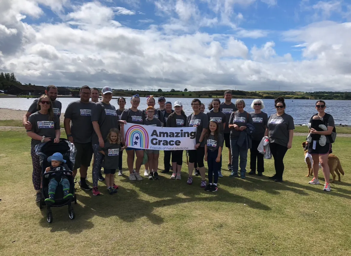 A group of people standing together, smiling and holding the Amazing Grace banner during a fundraising event. The backdrop features a serene lake, with clear blue skies and soft white clouds, creating a peaceful and uplifting atmosphere.