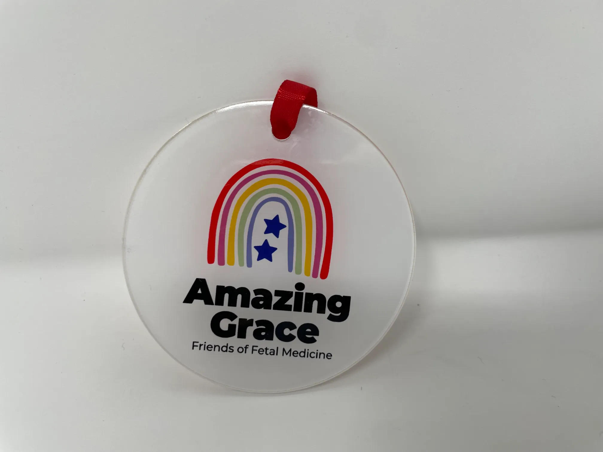 Flat plastic Amazing Grace round bauble decoration with logo and “Amazing Grace Friends of Fetal Medicine,” hanging with red ribbon