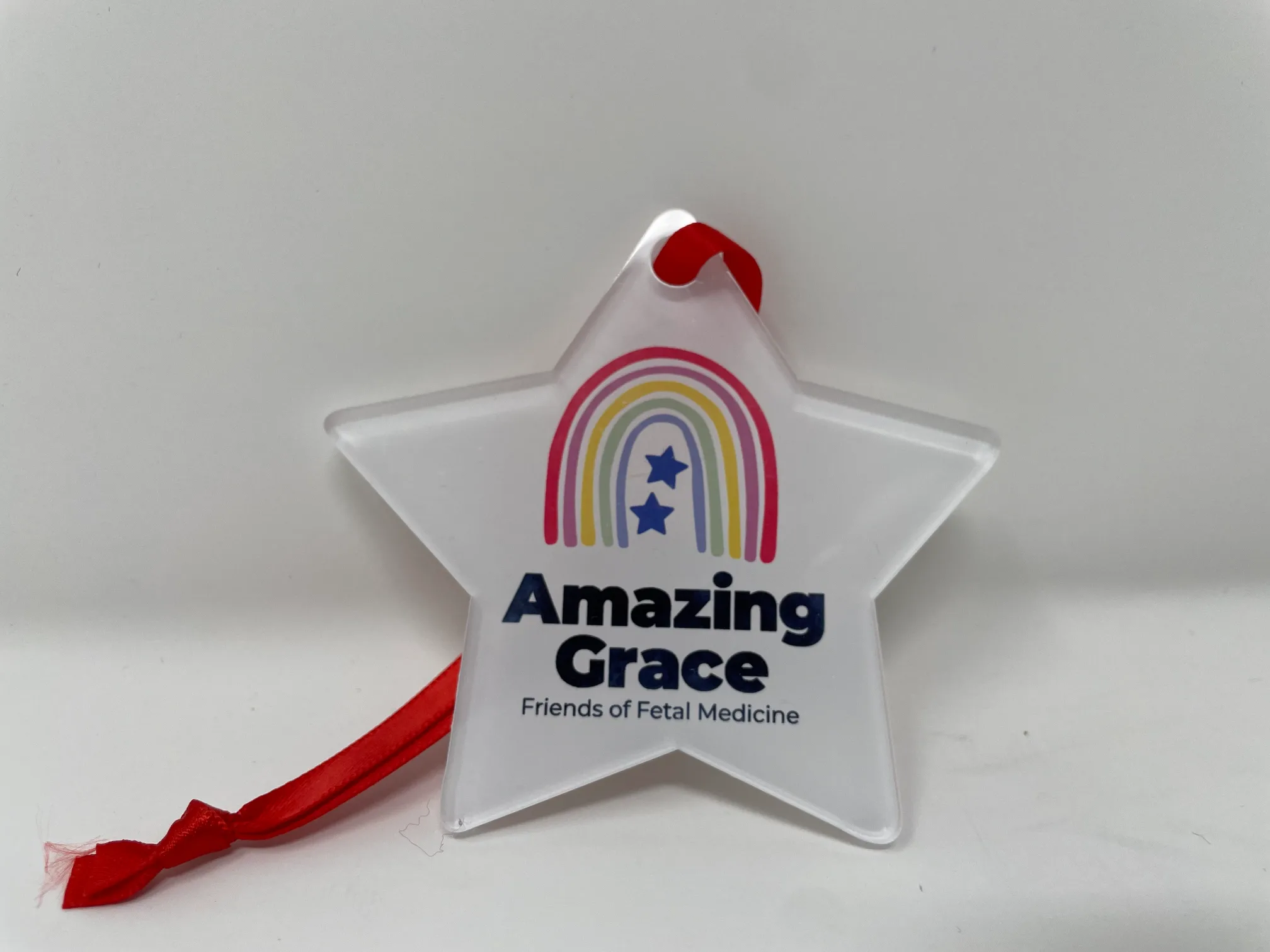 Flat plastic Amazing Grace Christmas star decoration with logo and “Amazing Grace Friends of Fetal Medicine,” hanging with red ribbon