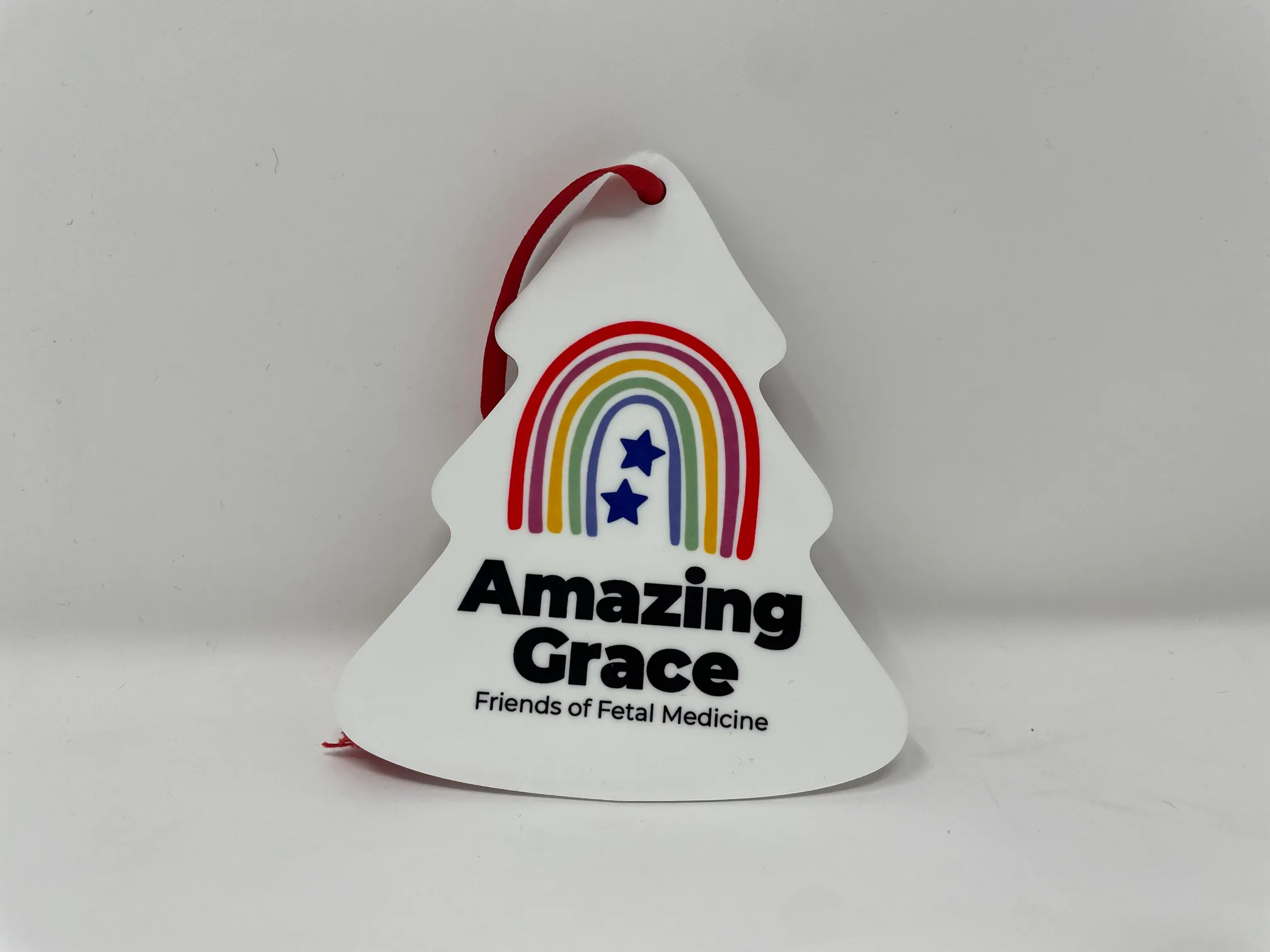 Flat plastic Amazing Grace Christmas tree decoration with logo and “Amazing Grace Friends of Fetal Medicine,” hanging with red ribbon